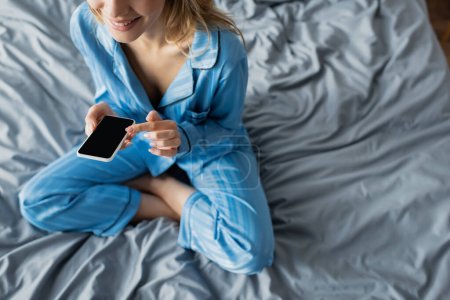 Foto de Vista recortada de la mujer sonriente en pijama azul usando un teléfono inteligente con pantalla en blanco mientras está sentada en la cama - Imagen libre de derechos