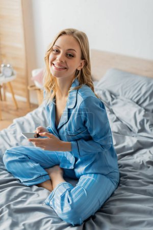 Photo for Positive woman in blue pajama holding smartphone with blank screen - Royalty Free Image