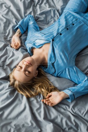 top view of carefree woman in blue silk pajama lying with closed eyes on bed in modern apartment 