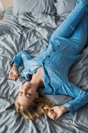 top view of pleased woman with tattoo lying with closed eyes on bed in modern apartment 