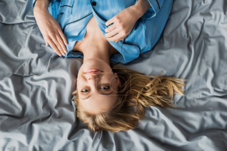 Draufsicht einer glücklichen Frau im blauen Seidenpyjama, die auf dem Bett liegt und in die Kamera blickt 
