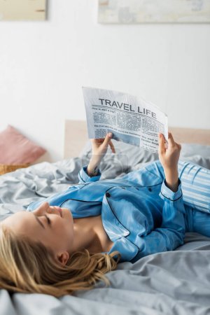 Photo for Tattooed young woman smiling while reading travel newspaper in bed - Royalty Free Image