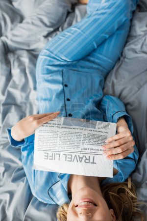 Foto de Vista superior de la mujer recortada y feliz sonriendo mientras lee la vida de viaje periódico en la cama - Imagen libre de derechos