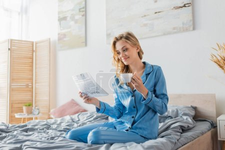 glückliche junge Frau liest Reisezeitung und hält Tasse Kaffee im Bett 