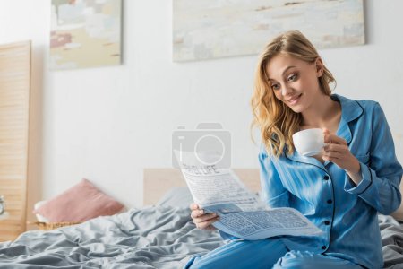 verblüffte junge Frau liest Reisezeitung und hält Tasse Kaffee im Bett 