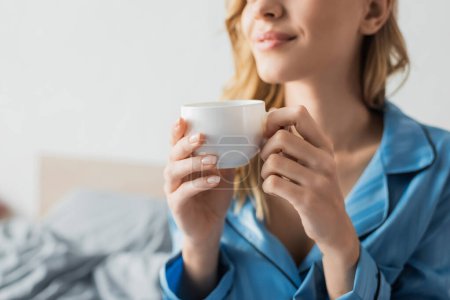 abgeschnittene Ansicht einer zufriedenen jungen Frau im blauen Pyjama mit einer Tasse Kaffee 