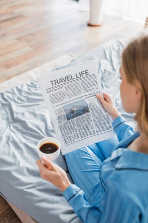 Foto de Vista aérea de la mujer rubia leyendo el periódico de la vida de viaje y sosteniendo la taza de café en el dormitorio - Imagen libre de derechos