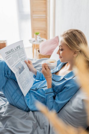 Téléchargez les photos : Vue de côté de jeune femme heureuse lecture Voyage journal de la vie et tenant une tasse de café au lit - en image libre de droit