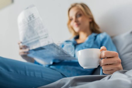 Photo for Cup of coffee in hand of young woman with newspaper on blurred background - Royalty Free Image
