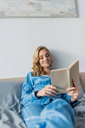 zufriedene junge Frau in blauer Seidennachtwäsche liest Buch im Schlafzimmer 