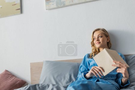 Photo for Pensive young woman in blue silk nightwear holding book in bedroom - Royalty Free Image