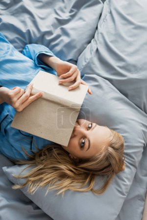 vista superior de la joven complacida en la cara de la cubierta de la ropa de dormir de seda azul con libro en la cama 