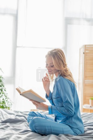 zufriedene junge Frau in blauer Seiden-Nachtwäsche liest Buch, während sie sich am Wochenende ausruht 