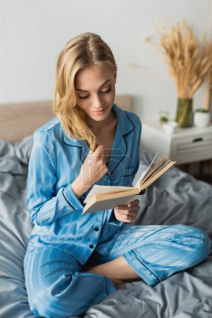 Blonde junge Frau in blauer Seiden-Nachtwäsche liest Buch, während sie am Wochenende ausruht 