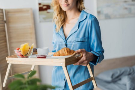 vista recortada de la mujer complacida en pijama azul bandeja de celebración con desayuno en el dormitorio 