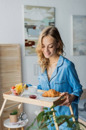 mujer feliz en pijama azul mirando bandeja con desayuno en el dormitorio 