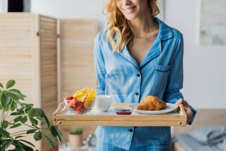 abgeschnittene Ansicht einer glücklichen Frau in blauer Nachtwäsche mit Tablett und Frühstück im Schlafzimmer 