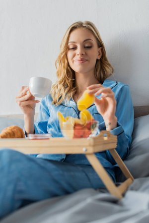 sonriente joven en pijama sosteniendo taza de café y mango seco cerca de la bandeja mientras desayunaba en la cama 