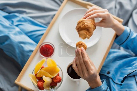 vista superior de la mujer recortada sosteniendo croissant fresco cerca de bandeja mientras desayuna en la cama 
