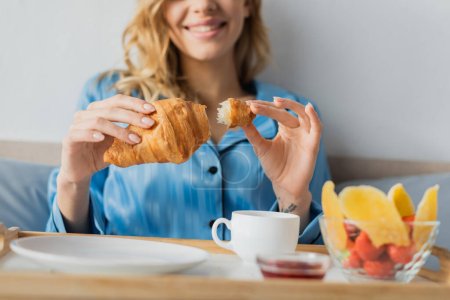 vista recortada de la joven sonriente en pijama sosteniendo croissant fresco cerca de bandeja con café mientras desayuna en la cama 