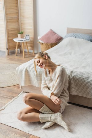Foto de Mujer rubia joven en suéter de punto y calcetines sentados en la alfombra en el dormitorio moderno - Imagen libre de derechos