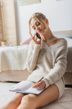 blonde woman in sweater talking on smartphone and taking notes in notebook 