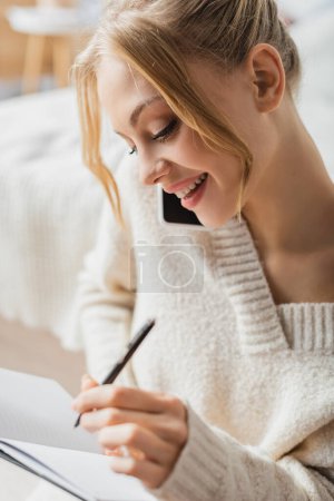 smiling woman in sweater talking on smartphone and taking notes in notebook 