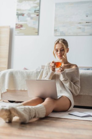 blonde woman holding glass cup of tea while looking at laptop in modern apartment 