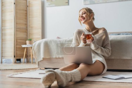 mujer feliz sosteniendo una taza de té mientras está sentada con un portátil en un apartamento moderno 