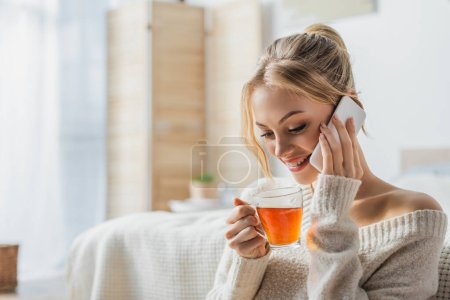 happy woman holding glass cup of tea while talking on smartphone in modern apartment 