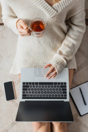 top view of cropped woman holding glass cup with tea while using laptop near smartphone with blank screen 