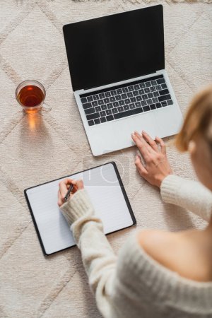 top view of woman taking notes near laptop and glass cup with tea on carpet 