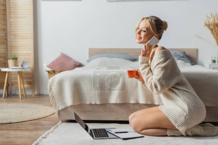 cheerful woman holding glass cup with tea and talking on smartphone near laptop on carpet 