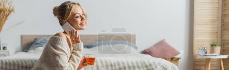 cheerful woman holding glass cup with tea and talking on smartphone in bedroom, banner 