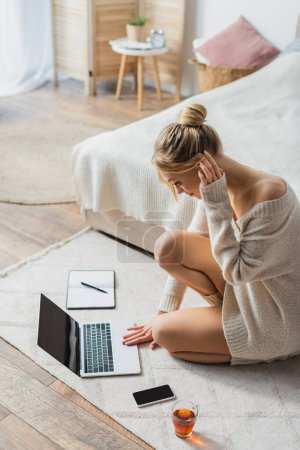 blonde woman in sweater using laptop near mobile phone and cup of tea on carpet in bedroom 