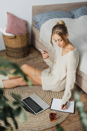 Foto de Mujer bonita en suéter tomando notas cerca de la computadora portátil y sosteniendo el teléfono inteligente mientras está sentado cerca de la cama - Imagen libre de derechos
