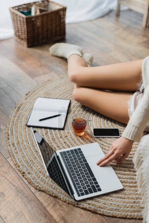 cropped view of young woman sitting near devices and cup of tea while working from home 