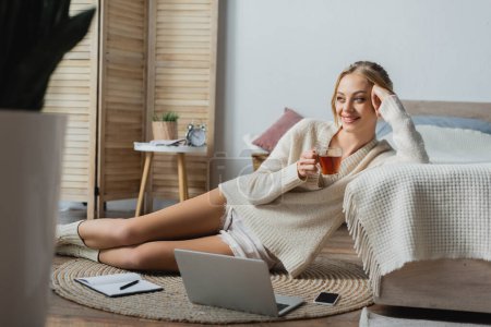smiling blonde woman in sweater holding glass cup with tea while sitting near gadgets in bedroom 