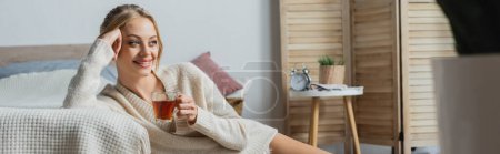 smiling blonde woman in sweater holding glass cup with tea in bedroom, banner 