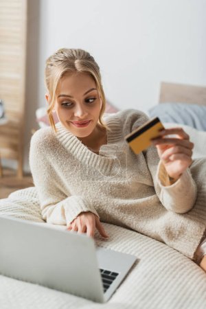 mujer alegre que sostiene la tarjeta de crédito y el uso de la computadora portátil mientras que hace compras en línea en dormitorio 