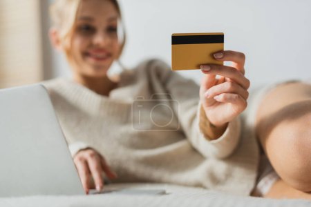verschwommene Frau mit Kreditkarte und Laptop beim Online-Shopping im Schlafzimmer 