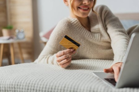 Téléchargez les photos : Vue partielle de femme gaie tenant la carte de crédit et utilisant un ordinateur portable tout en faisant des achats en ligne dans la chambre - en image libre de droit