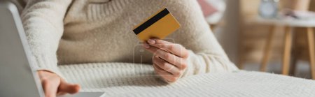 vista parcial de la mujer que sostiene la tarjeta de crédito y el uso de la computadora portátil mientras hace compras en línea en el dormitorio, bandera 