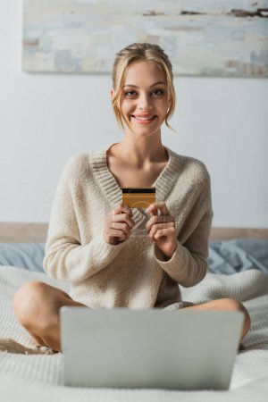 cheerful woman holding credit card near laptop while doing online shopping in bedroom 