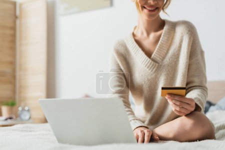 Teilbild einer glücklichen Frau, die beim Online-Shopping im Schlafzimmer ihre Kreditkarte in der Nähe des Laptops hält 