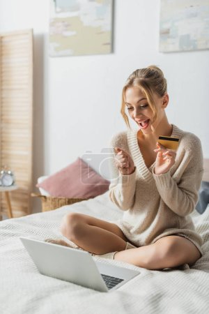 Erstaunte Frau mit Kreditkarte in der Nähe von Laptop beim Online-Shopping im Schlafzimmer 