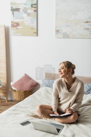 happy blonde woman taking notes near gadgets on bed in modern apartment 