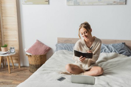 cheerful blonde woman taking notes near gadgets on bed in modern apartment 