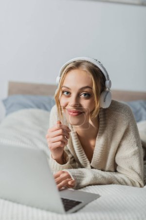 happy young woman in wireless headphones looking at camera while lying near laptop in bedroom 