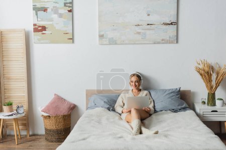 Foto de Longitud completa de la joven alegre en auriculares inalámbricos utilizando el ordenador portátil en el dormitorio moderno - Imagen libre de derechos
